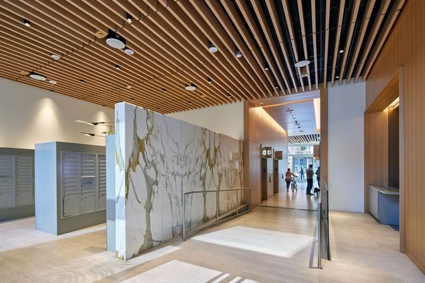 A bright contemporary commercial lobby or building entry with a prominent veined stone privacy screen, pale stone flooring, timber slatted ceilings, integrated recessed lighting, timber wall panelling, service cabinetry, and a corridor extending into the background. Mailboxes or locker panels are visible on the left, while people are present in the distant hallway.