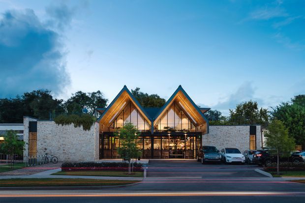 Exterior architectural photograph of a contemporary low-rise commercial building at dusk. The facade combines pale masonry walls with two prominent, steeply pitched timber-clad gabled roof forms, extensive glazed entrances, and dark structural framing. A landscaped forecourt and parking area sit in front of the building, with mature trees behind it.