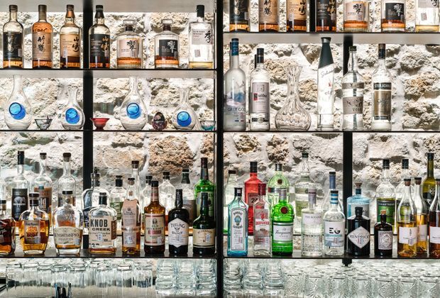 A close-up view of a backlit bar display featuring multiple rows of liquor bottles, decanters, and glassware arranged on dark metal-framed shelving. The shelving is set against a rough exposed stone wall, creating a dramatic, richly textured bar backdrop. The image focuses on the built-in display and bar storage rather than a full room.