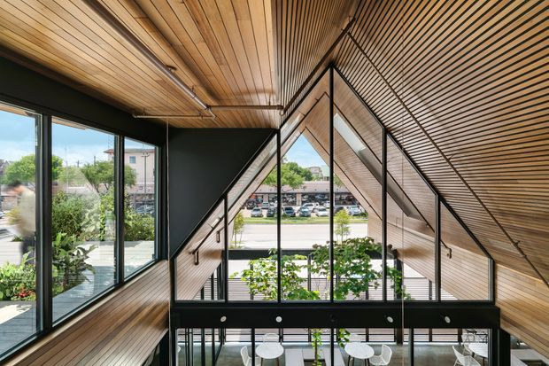 A dramatic contemporary interior featuring a sculptural black metal staircase and glazed stairwell rising through a double-height space. Warm timber slatted cladding covers the ceiling and angled walls, while extensive floor-to-ceiling glazing brings in views of the surrounding urban environment and greenery. A dining area is visible below the stair landing.
