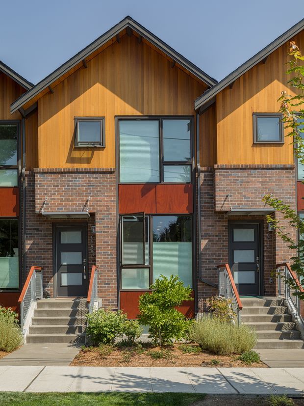Front exterior view of a contemporary multi-unit townhouse development. The facade combines warm vertical timber cladding, exposed reddish-brown brickwork, dark-framed windows and doors, and steeply pitched gabled roofs. Individual entries are reached by concrete steps with metal handrails, with low-maintenance landscaped planting and a sidewalk in front.