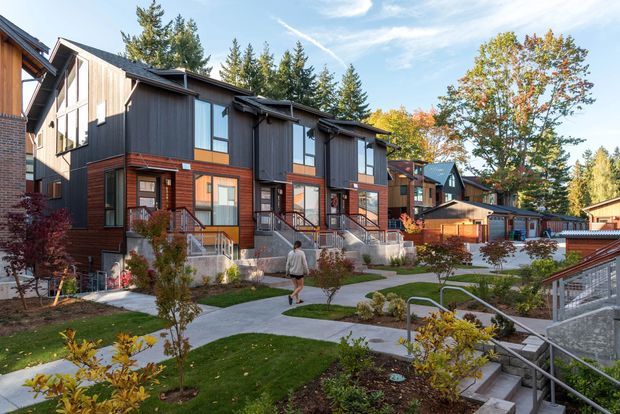 A landscaped residential townhouse or apartment complex with multiple attached multi-storey homes arranged along curved concrete walkways. The buildings feature dark vertical cladding, warm reddish-brown timber or painted siding, brick accents, pitched roofs, large windows, and individual front entries with stairs and metal handrails. Young planted gardens and lawns surround the pedestrian paths, with mature trees in the background under a bright sky.