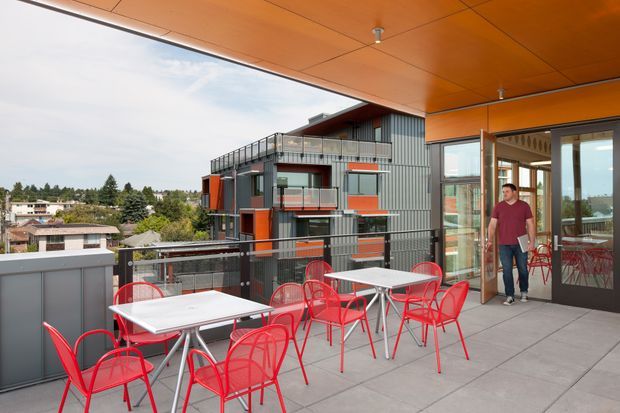 A contemporary rooftop or upper-level outdoor terrace with grey tiled paving, bright red metal café chairs, white tables, and a compact outdoor kitchen or service cabinet. An orange soffit and structural beam frame the terrace, which opens through large glazed doors to an interior dining area. Across the street is a modern multi-level building with corrugated metal cladding and balconies. The terrace overlooks a suburban neighbourhood and appears designed for casual outdoor dining and entertaining.