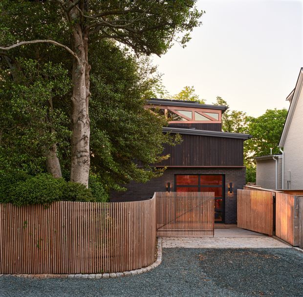 A contemporary dark-clad residential house viewed from the street, set behind a curved vertical timber-slat fence and gate. The compact two-storey facade features dark vertical timber cladding, exposed warm timber detailing, red-brown framed glazing, and a low sloping roof. A gravel driveway and paved entry lead to the front door, with mature trees and planting screening the property.