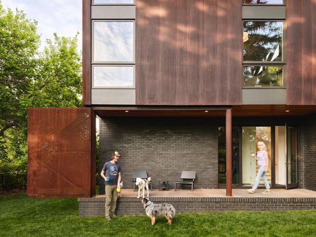 Exterior view of a contemporary multi-storey home set among mature trees. The facade combines dark brick at the lower level with warm rust-toned metal cladding above and a projecting upper floor. Large rectangular windows, a covered outdoor terrace, slim round support columns, and a broad pivoting metal gate or screen are visible. The image also shows lawn, trees, people, and dogs.