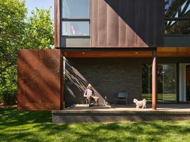 Exterior view of a contemporary multi-level home with a covered outdoor sitting area beneath the upper floor. The facade combines dark timber cladding, dark brick, large black-framed glazing, and a rust-toned perforated metal screen. A raised brick-edged deck, outdoor chairs, a seated person, and a dog are visible, with lawn and mature trees surrounding the house.