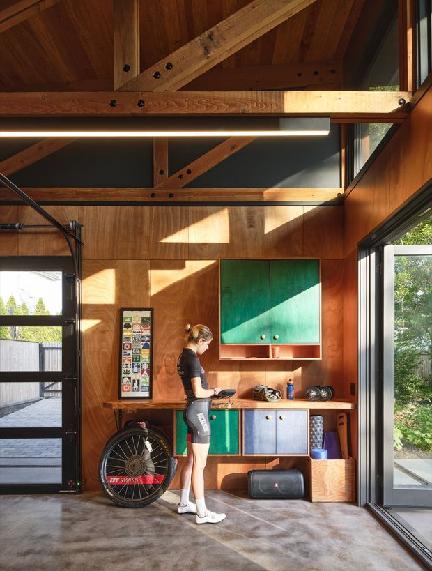 A compact, sunlit multipurpose garage or studio interior with exposed timber framing and warm plywood wall and ceiling lining. Large glazed garage doors and windows open to the outdoors. A colourful built-in workbench and storage cabinetry in teal, blue, and natural timber occupies the rear wall, with a stationary exercise bike, sports equipment, and a person using the workspace. The space has an industrial-workshop character softened by custom joinery and abundant natural light.
