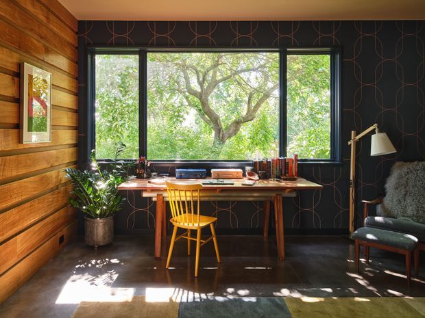 A warm, characterful home office arranged beside large black-framed windows overlooking leafy greenery. The room features horizontal timber wall panelling, dark patterned wallpaper, a compact wooden desk with books and accessories, a bright yellow spindle-back chair, a floor lamp, and a dark upholstered armchair. Strong natural light creates a cosy yet vibrant workspace atmosphere.