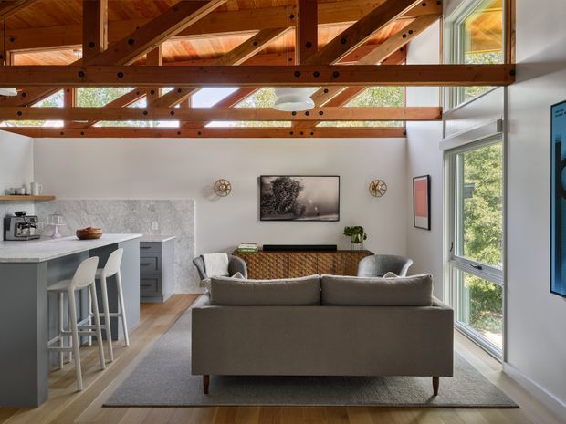 A compact open-plan living room and kitchen with a contemporary fit-out. The room features a grey sofa facing a media console, grey upholstered lounge chairs, a light grey area rug, timber flooring, a pale stone-look kitchen benchtop and splashback, and white walls. Large windows provide abundant natural light and views of greenery. Exposed warm timber roof trusses and beams create a strong structural feature overhead.