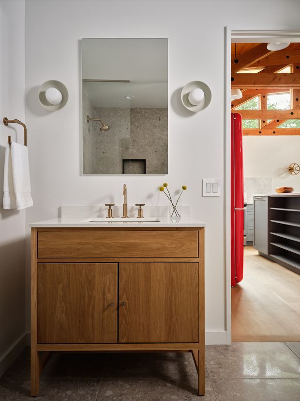A contemporary bathroom vanity area with a freestanding walnut-toned timber vanity, white countertop, brass tapware, round wall sconces, rectangular mirror, and light grey stone-look tiled surfaces. Through the open doorway, part of an adjacent kitchen or utility space is visible, featuring exposed timber beams, pale cabinetry, open shelving, and a bright red refrigerator.
