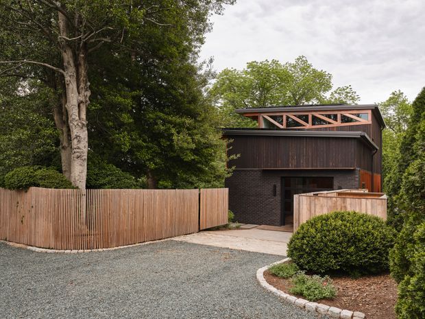 Exterior view of a contemporary dark-clad residential building set within a leafy landscaped site. The compact structure features vertical dark timber cladding, a black brick lower level, a projecting roof form, and a high clerestory-like band with exposed pink-toned timber diagonal bracing. A timber privacy fence, gravel forecourt, concrete entry path, clipped shrubs, and mature trees frame the building.