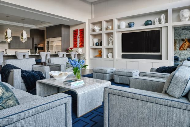 A bright, refined open-plan living room with multiple upholstered armchairs and sofas arranged around a pale central ottoman-style coffee table. Built-in white shelving surrounds a large wall-mounted television, while the adjoining kitchen features dark timber cabinetry, a light island bench, pendant lighting, and bar seating. Blue and grey furnishings, patterned rug, decorative objects, and floral accents create a polished contemporary interior.