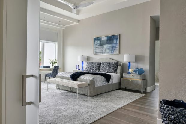 A bright, refined bedroom viewed through an open doorway. The room features a large upholstered bed with patterned grey bedding and cushions, matching bedside tables with lamps, a long upholstered bench, a large pale area rug, timber-look flooring, light neutral walls, recessed ceiling detailing, and a ceiling fan. Blue accents and artwork add contrast. A small desk or sitting area is visible near the window.