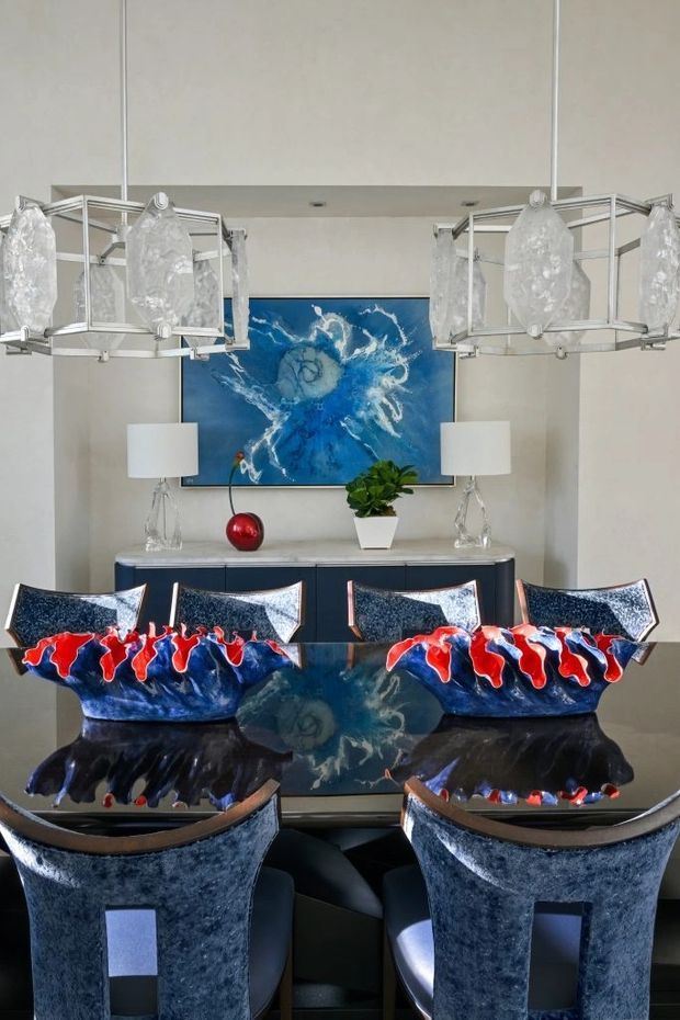 A refined dining room centered on a glossy dark dining table with upholstered dining chairs, a dark sideboard, paired table lamps, and dramatic sculptural red-and-blue ceramic vessels. A large abstract blue artwork hangs on the pale wall, while two oversized geometric pendant lights with translucent sculptural shades frame the composition.