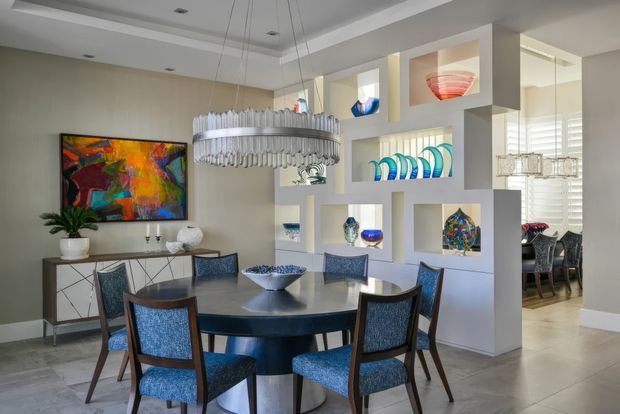 A refined dining room featuring a round dark dining table with blue upholstered chairs, a large abstract artwork, a sculptural suspended chandelier, and a white geometric open display partition with colourful glass and ceramic objects. A mirrored wall reflects additional dining furniture and glazing, while recessed ceiling lighting and neutral finishes create a polished contemporary interior.