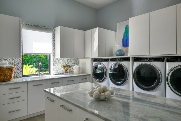 Bright, spacious laundry room with extensive white built-in cabinetry, a large central island with a pale grey veined stone benchtop, patterned mosaic tile splashback, and multiple front-loading washing machines and dryers. A window with a roller blind brings in natural light, while cool grey walls and white joinery create a clean, contemporary utility space.