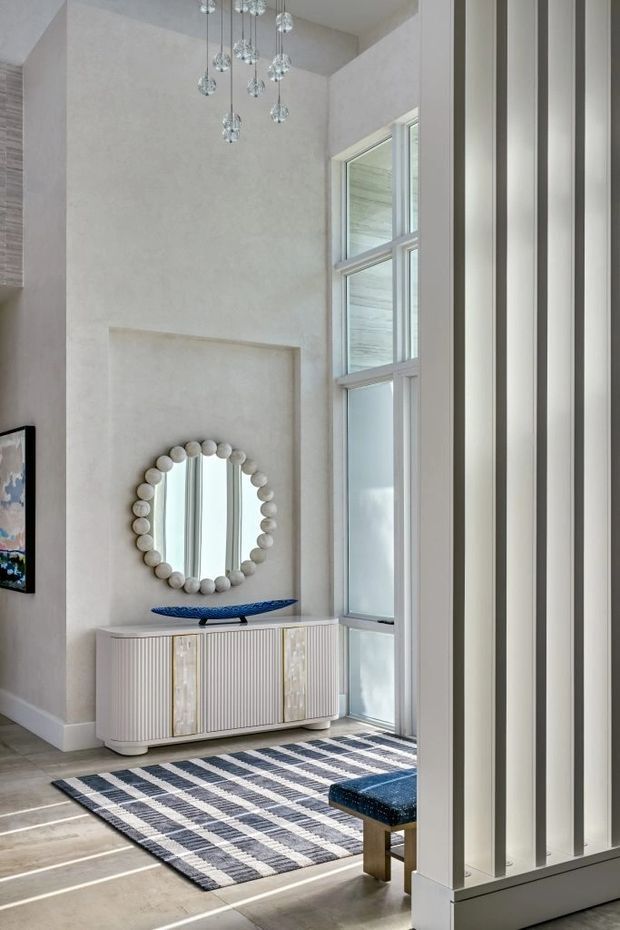 A tall, light-filled contemporary entry hall with double-height proportions, pale textured walls, large vertical windows, and a sculptural cluster pendant. A fluted white sideboard with decorative doors sits beneath a round bead-framed mirror, with a blue vessel on top. A navy, white, and grey patterned rug defines the limestone- or stone-floored space, while vertical white architectural screening frames the right side.