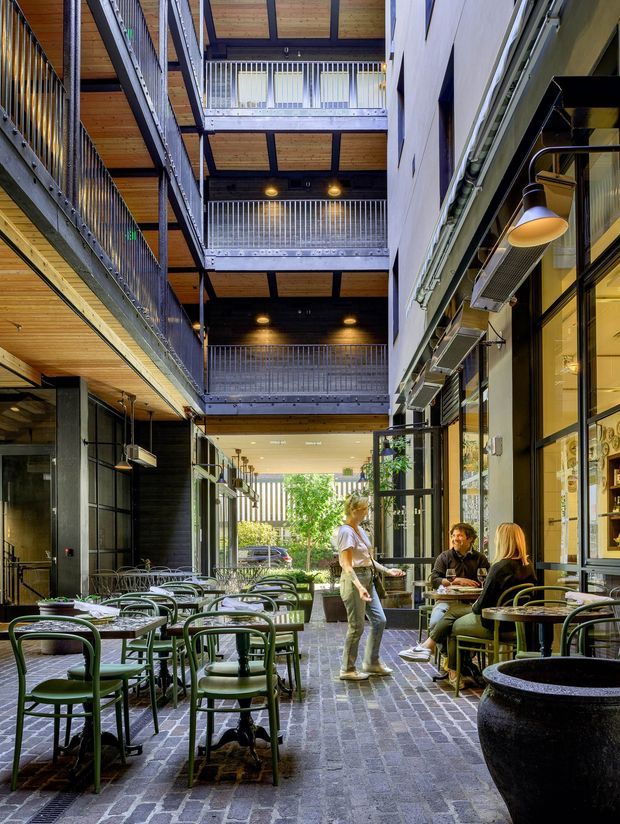 A covered multi-level urban laneway or courtyard café with outdoor dining tables and green chairs, dark brick paving, timber-lined ceilings, exposed structural steel, and stacked balconies with metal balustrades. Retail or hospitality shopfronts line both sides, with diners seated near the entrance and daylight opening toward a planted street edge.