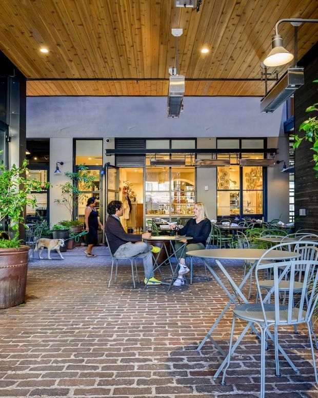 A covered outdoor café or commercial courtyard with exposed timber decking overhead, exposed mechanical services, dark steel framing, brick paving, café tables and chairs, planting, and a glazed storefront or dining area in the background. The scene has an industrial, lively hospitality character with warm interior lighting.