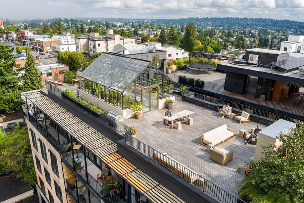 Aerial view of a large rooftop terrace in a dense urban neighbourhood. The terrace has timber decking, outdoor lounge furniture, planters, railings, and a prominent glass greenhouse or conservatory structure. Adjacent rooftop structures include dark cladding, solar panels, and rooftop equipment, with surrounding residential and commercial buildings visible in the background.