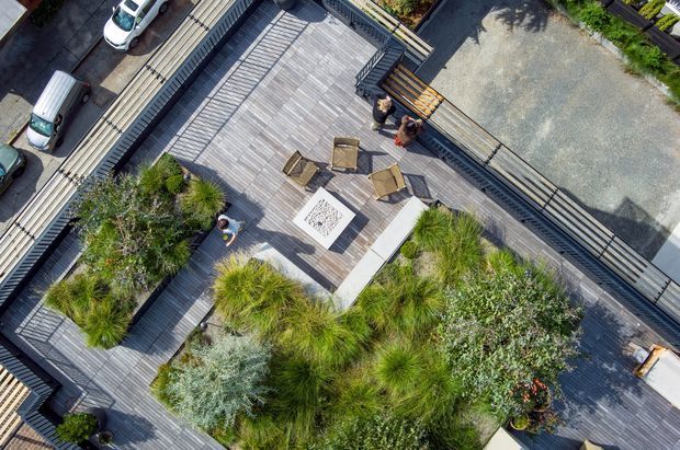 Aerial view of a landscaped rooftop terrace or elevated outdoor deck, featuring extensive timber-look decking, planted garden beds with ornamental grasses and shrubs, a central outdoor fireplace or fire pit, lounge chairs, and dark metal or timber privacy railings. Adjacent driveway and parked vehicles are visible at the edges.