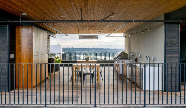 Open-air rooftop or upper-level terrace with an outdoor kitchen and dining area. The space features a warm timber-planked ceiling and decking, exposed black metal services and railings, built-in cabinetry, a barbecue grill, bar-height dining table and stools, and expansive views beyond the balustrade. The design has a refined industrial character with strong indoor-outdoor connection.