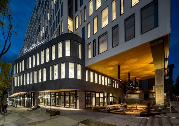 Dusk exterior view of a multi-storey contemporary commercial building with a curved, glazed corner frontage, dark framed windows, illuminated interiors, and a large timber-lined cantilevered entrance canopy. The active street-level frontage includes extensive glazing, outdoor bench seating, and a covered entry plaza supported by substantial stone-clad columns.