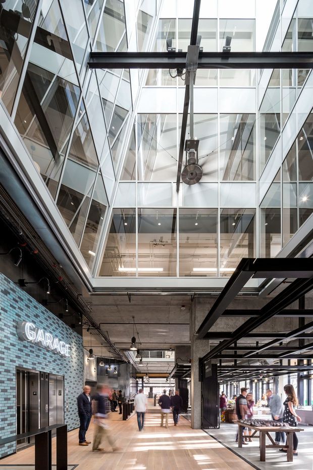 A tall, multi-level commercial interior atrium with extensive glazed walls, exposed concrete ceilings, black steel structural framing, visible services, and an overhead suspended industrial mechanism. The ground level is a bright public circulation and gathering space with elevators, long tables, seating, people, and a prominent illuminated “GARAGE” sign. The architecture combines a modern glass enclosure with industrial warehouse-style detailing.