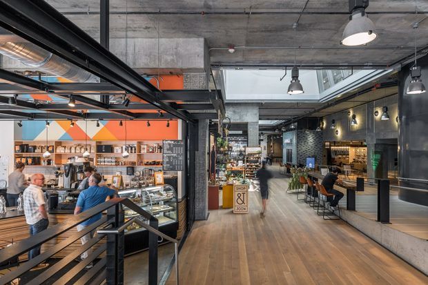A spacious industrial-style café and hospitality interior viewed from a stair landing. The open-plan space features exposed concrete ceilings, black metal framing and ductwork, timber flooring, pendant and wall lighting, a café counter with display cases, bar seating, retail shelving, and a skylight. Customers and staff occupy the café area.