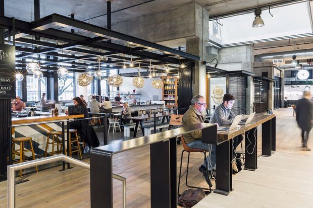 A spacious, industrial-style café or hospitality venue with exposed concrete ceilings and columns, black steel framing, open metal shelving, timber floors, bar-height communal tables, and a service counter. Pendant globe lights and track lighting illuminate the dining and coworking areas, with large glazed openings bringing in abundant daylight. Several patrons are seated at tables and counters, and a laptop workspace is visible in the foreground.