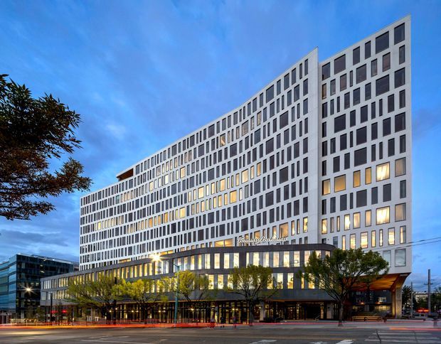 Exterior architectural photograph of a large contemporary multi-storey urban building, likely a hotel or mixed-use development. The facade is composed of repetitive white rectangular window bays with stepped massing, while a darker glazed podium with illuminated ground-floor commercial or lobby spaces anchors the street frontage. Photographed at dusk with illuminated interiors, street trees, traffic lanes, and a blue evening sky.