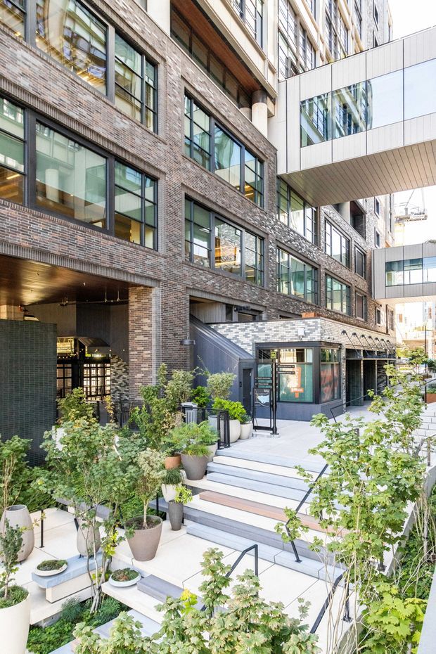 A landscaped urban courtyard and building entrance framed by multi-storey brick and glass facades. Broad pale concrete steps with dark metal handrails lead to a recessed entrance and a small glazed pavilion-like reception or lobby structure. An enclosed bridge spans between the buildings overhead. The architecture combines textured multi-toned brickwork, large dark-framed windows, light metal-clad volumes, and timber-lined soffits, with abundant potted and planted greenery throughout.