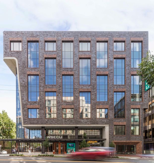 Street-level photograph of a mid-rise urban commercial building with a dark reddish-brown brick facade, strong vertical window bays, extensive reflective glazing, and a sheltered ground-floor entrance. Adjacent buildings, trees, pedestrians, and passing traffic are visible in the streetscape.