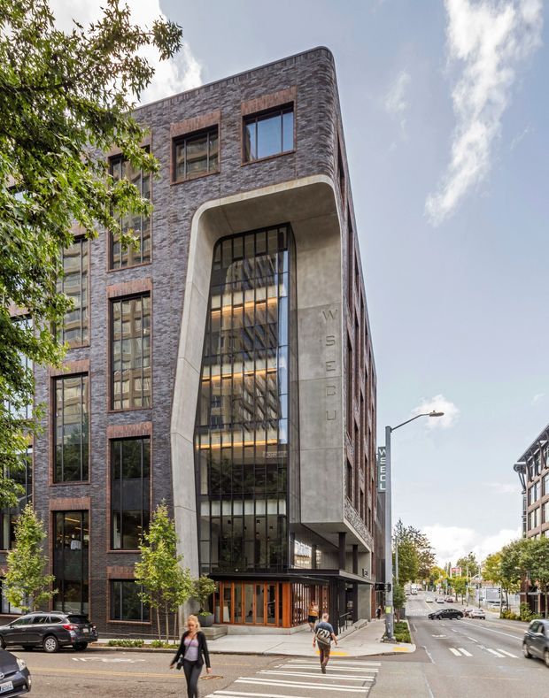 Street-level exterior photograph of a contemporary multi-storey commercial building on an urban corner. The facade combines dark textured brick with large vertically proportioned windows, warm timber-framed detailing, and a prominent curved concrete entrance surround with tall black-framed glazing. Pedestrians, street trees, road markings, and neighbouring buildings reinforce the dense urban setting.