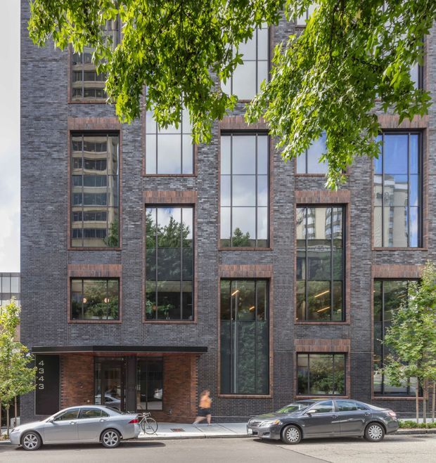 Street-level view of a contemporary multi-storey urban building with a dark grey-brown brick facade, strong vertical window openings, and contrasting reddish brick window surrounds. The ground floor includes a recessed glazed entry, with mature street trees and parked cars in the foreground.