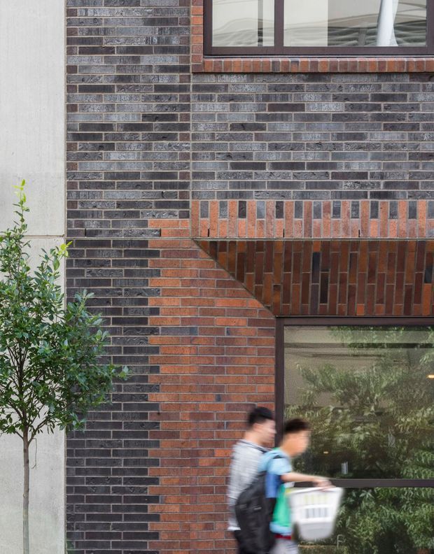 Close-up exterior architectural view of a contemporary brick facade combining dark charcoal brick with warm red-orange brick detailing. The wall features carefully patterned brickwork, projecting and angled courses, dark-framed windows, and a large glazed opening. A small tree and blurred pedestrians appear in the foreground.
