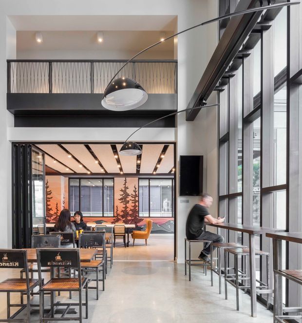 A contemporary multi-level commercial interior with a café or hospitality seating area. The space features polished concrete flooring, tall black-framed windows, an open mezzanine with patterned metal balustrades, exposed dark structural elements, and large curved pendant lights. Café tables, stools, and chairs occupy the foreground, while a colourful mural and additional seating are visible deeper in the space.