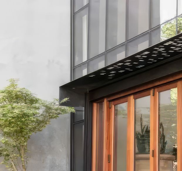 Close-up exterior view of a contemporary building entrance, featuring dark grey rendered walls, tall translucent or glazed upper facade panels, a black perforated projecting canopy, and warm timber-framed glazed doors. A leafy tree partially frames the left side.
