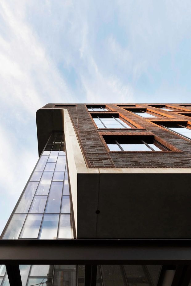 Low-angle view looking up at a multi-storey urban building facade. The facade combines dark reddish-brown brickwork, projecting window surrounds, extensive glazed vertical sections, and dark cantilevered or recessed slab elements. The composition emphasizes the building's height and strong geometric lines against a pale blue sky.