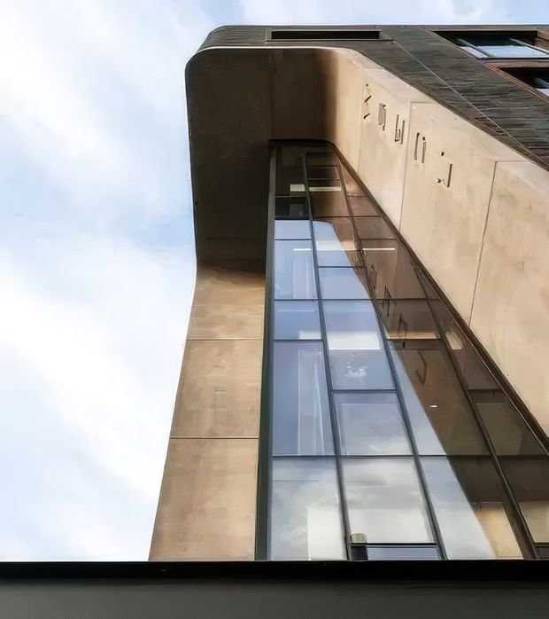Upward-looking architectural photograph of a tall contemporary building facade. The composition is dominated by a vertical glazed curtain wall framed in dark metal, bordered by smooth warm-toned masonry or rendered wall surfaces. A curved projecting roof or canopy edge is visible near the top, with additional dark cladding and rooftop detailing. The sky is visible around the building, emphasizing its height and vertical perspective.