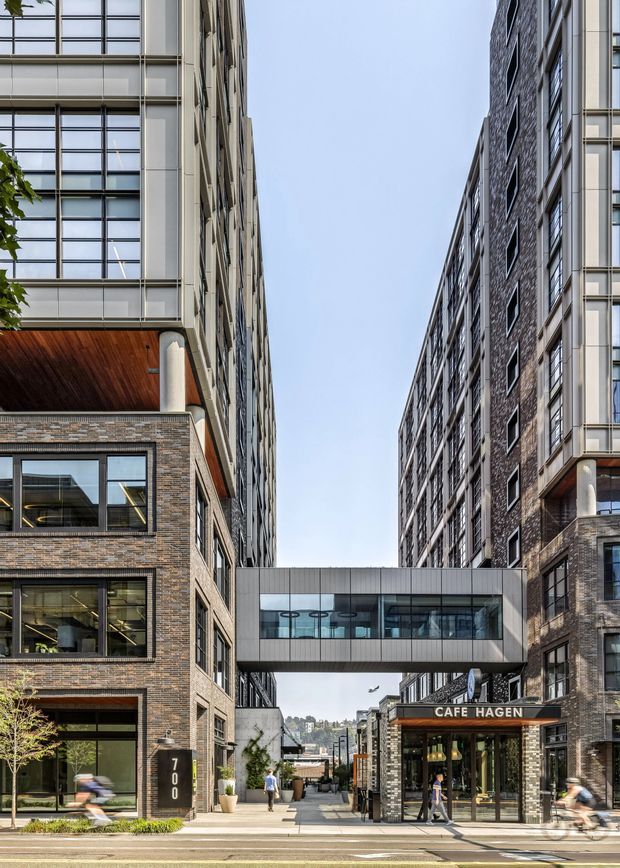 Street-level view of a contemporary multi-storey urban development with brick and light metal-clad facades, extensive grid-pattern glazing, and a glazed pedestrian skybridge connecting two buildings. Ground-floor commercial tenancy includes a Café Hagen storefront, with a paved pedestrian passage and distant hillside urban context visible beyond.