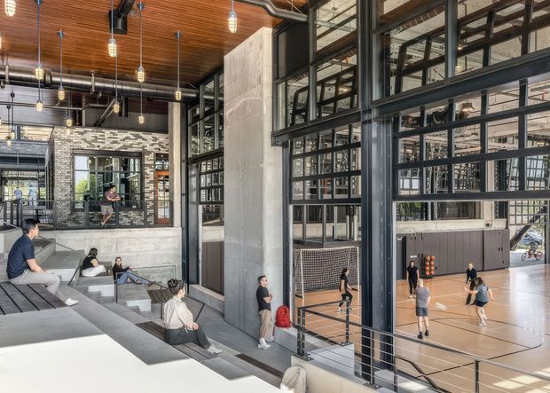 Interior view of a large contemporary industrial-style sports and recreation facility. A full-height glazed steel partition overlooks an indoor basketball court, while tiered concrete seating occupies the foreground. The space features exposed concrete columns, dark metal framing, exposed ductwork, a warm timber slatted ceiling, and suspended pendant lights.