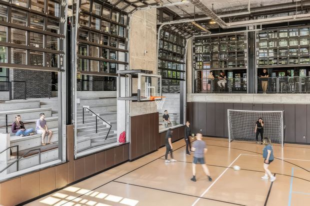 A bright multi-level indoor sports hall with a polished timber court, basketball hoop, futsal goals, and players. Large industrial-style glazed walls and exposed structural services surround the court, with tiered concrete seating and an upper-level balcony visible. The space has a contemporary institutional or commercial character with abundant natural light.