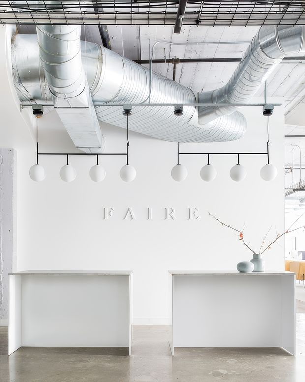 Bright minimalist commercial reception or retail interior with a white feature wall embossed with the word “RAIRE,” two simple white reception counters, sculptural white globe pendant lights, exposed silver HVAC ductwork, and a polished concrete floor. The space has an open industrial ceiling and a clean, gallery-like aesthetic.