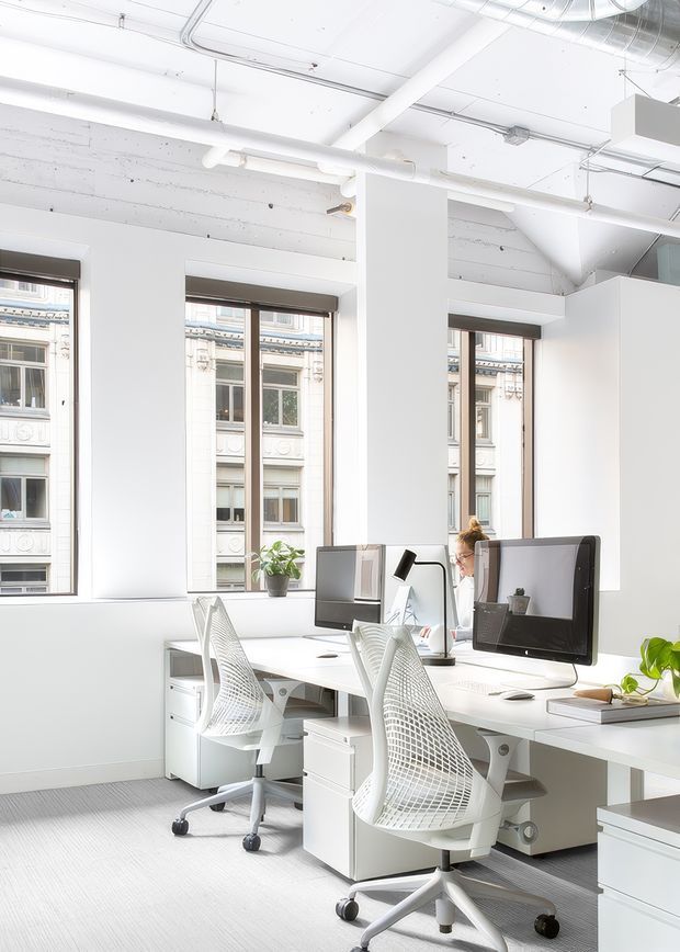 Bright open-plan office interior with rows of white desks, computer monitors, ergonomic mesh office chairs, potted plants, and large windows overlooking an ornate urban building. The space has exposed white-painted services and ductwork, creating a clean, contemporary commercial workplace.