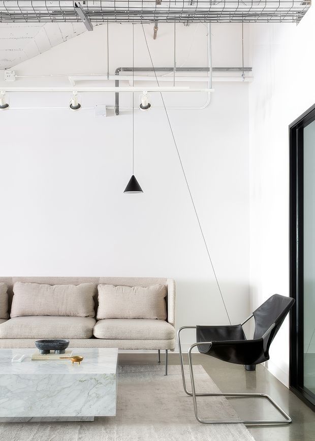 Bright minimalist living room with a low cream upholstered sofa, black leather sling chair, pale marble coffee table, and neutral rug. White walls and a white-painted exposed ceiling with visible services, track lighting, and a suspended black pendant create a refined contemporary loft-like interior. Full-height black-framed glazing is visible on the right.