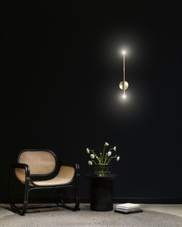 A moody, minimalist interior vignette with a black wall, a sculptural glowing pendant light, a black-framed woven cane lounge chair, and a small dark side table holding a glass vase of white flowers. Books rest on a textured neutral rug over a dark floor, creating a refined, high-contrast setting.