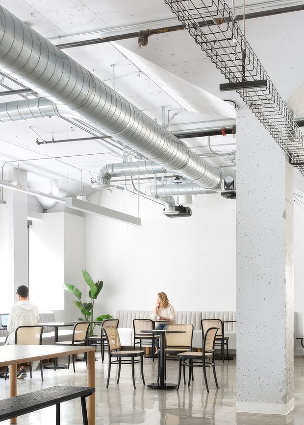 Bright contemporary commercial interior resembling a café, coworking space, or casual office lounge. The high white ceiling exposes extensive silver HVAC ductwork, suspended linear lighting, cable trays, and other building services. White walls and a large speckled structural concrete column frame a polished light floor. Small café-style tables and black-framed cane or upholstered chairs surround a long built-in bench, with a few people seated and a potted plant adding greenery.