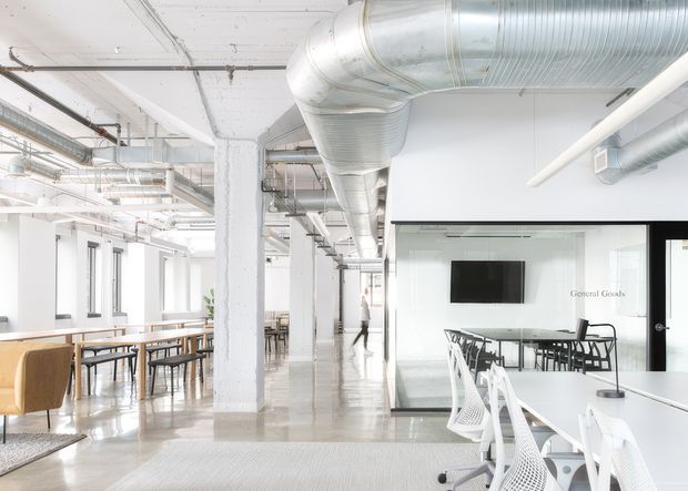 Bright open-plan commercial interior with exposed white structural columns, polished concrete flooring, extensive exposed silver HVAC ductwork, rows of desks and dining-style tables, white mesh office chairs, and a glazed meeting room with a wall-mounted display. The space has an airy, contemporary industrial office fit-out.