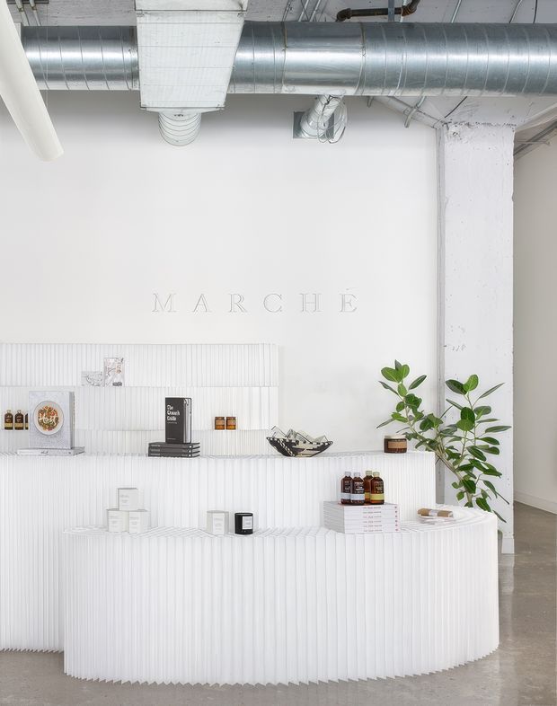 A minimalist commercial retail or reception space with a sculptural white fluted counter and matching fluted wall display. The bright white interior features exposed industrial ductwork and a painted structural column, subtle wall lettering reading “MARCHÉ,” curated product displays, and a large indoor plant. The floor is a smooth grey concrete finish, creating a clean, refined industrial atmosphere.